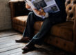 Man reading the newspaper on the a brown couch.