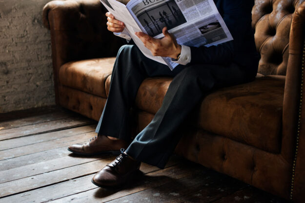 Man reading the newspaper on the a brown couch.